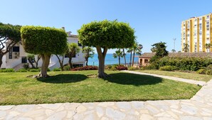 A landscaped area with two neatly trimmed trees, a stone pathway, and a view of the sea and buildings in the background. - Charming Apartment with Sea Views & Wi-Fi (Mijas Costa)