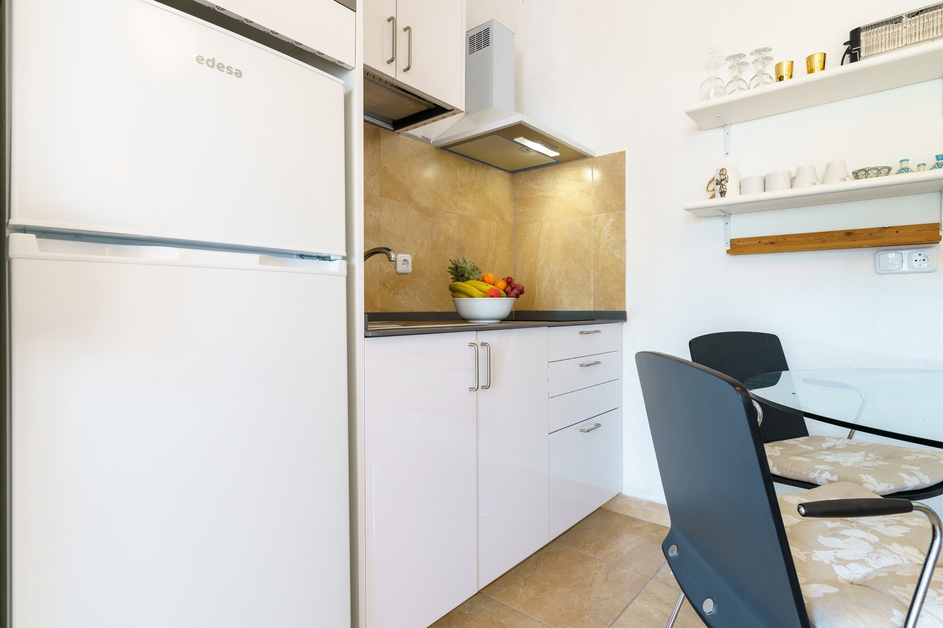 Cuina amb armaris blancs, un plat de fruita sobre el taulell i una zona de menjador amb una cadira.