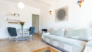 A modern living room with a sofa, chairs, a coffee table with seashells, a dining area with chairs and a table, and a kitchen area with shelves and a vase of flowers. - Charming Apartment with Sea Views & Wi-Fi (Mijas Costa)