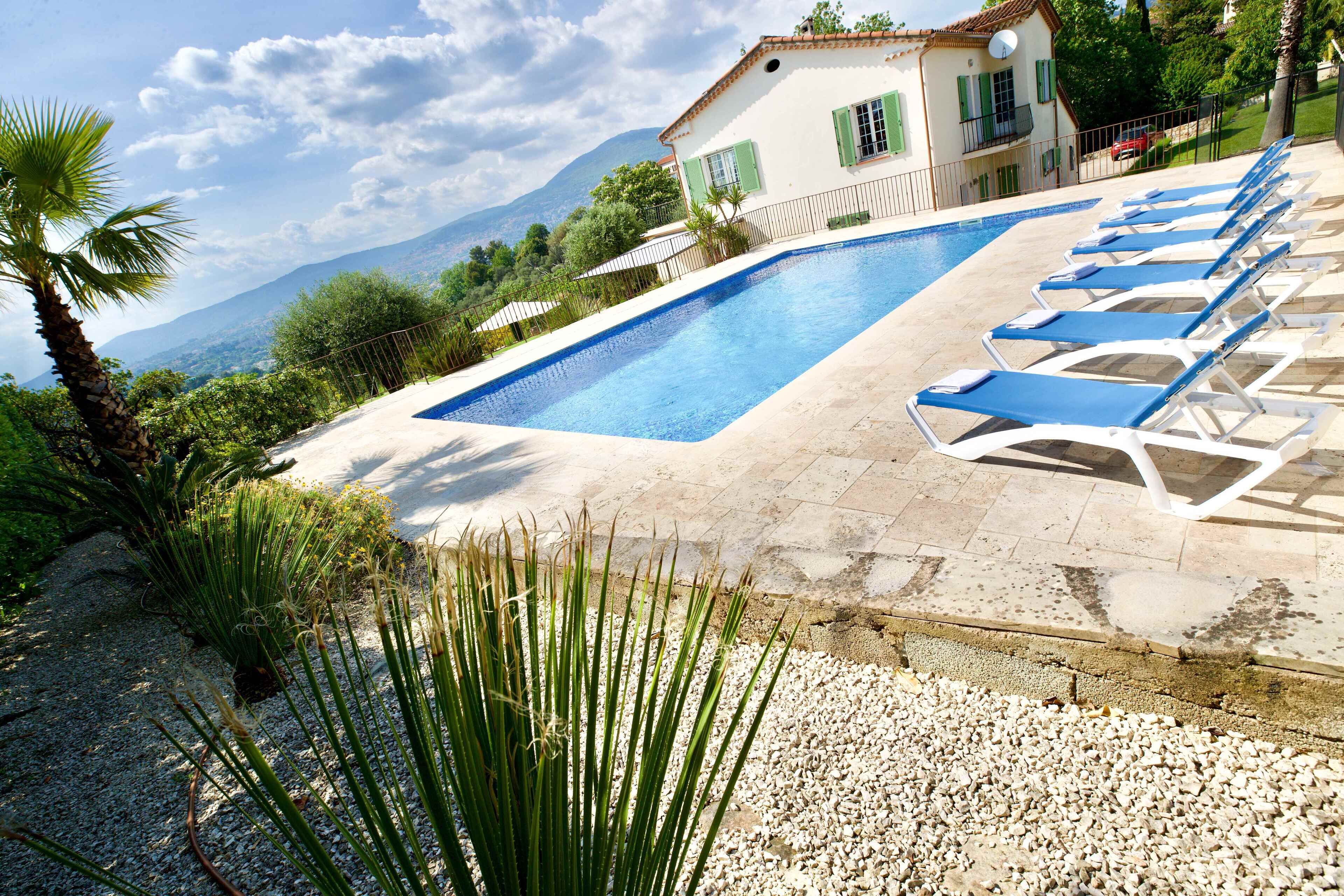 Ein Swimmingpool mit Liegestühlen, ein Haus mit Balkon und eine Bergkulisse im Hintergrund.