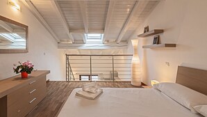 Ein Schlafzimmer mit Dachfenster, Holzbalken an der Decke, einem Bett mit weißen Leinen, einer Holzkommode und einer Topfpflanze.