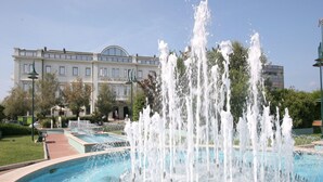 A fountain with multiple water jets in a circular pool, surrounded by a well-maintained garden and a building with a large arched window in the background. - Escape in Central Cattolica (Cattolica)