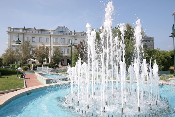 Une fontaine dotée de plusieurs jets d’eau, située dans un bassin circulaire, et entourée d’un jardin bien entretenu. Un bâtiment avec une grande fenêtre en arc de cercle se trouve en arrière-plan.