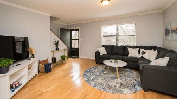Una sala de estar con un sofá negro, una mesa de centro blanca, un televisor sobre un mueble blanco y una escalera que lleva a otra habitación.