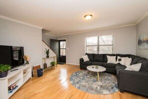 A living room with a black sofa, a white coffee table, a TV on a white stand, and a staircase leading to another room. - Great Deal! Walkable Condo Near Greenway + Mission (Asheville)