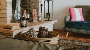 A cozy living room with a fireplace, a wooden table with a bottle of wine, a candle, and a decorative item. There is a sofa with a patterned cushion and a rug on the floor. - AGRITURISMO LA PAPESSA (Barberino Tavarnelle)