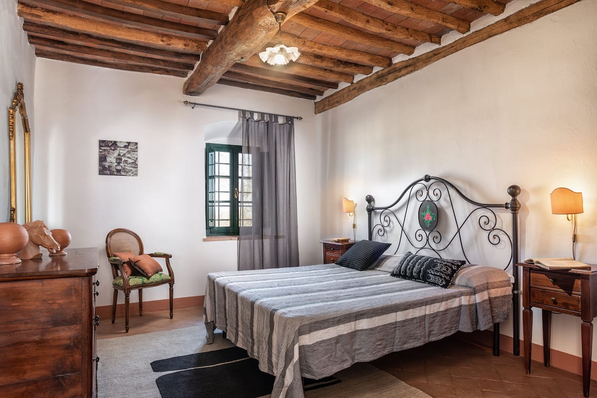 A bedroom with a wooden ceiling, a bed with a striped bedspread, two bedside tables with lamps, a chair, and a dresser.