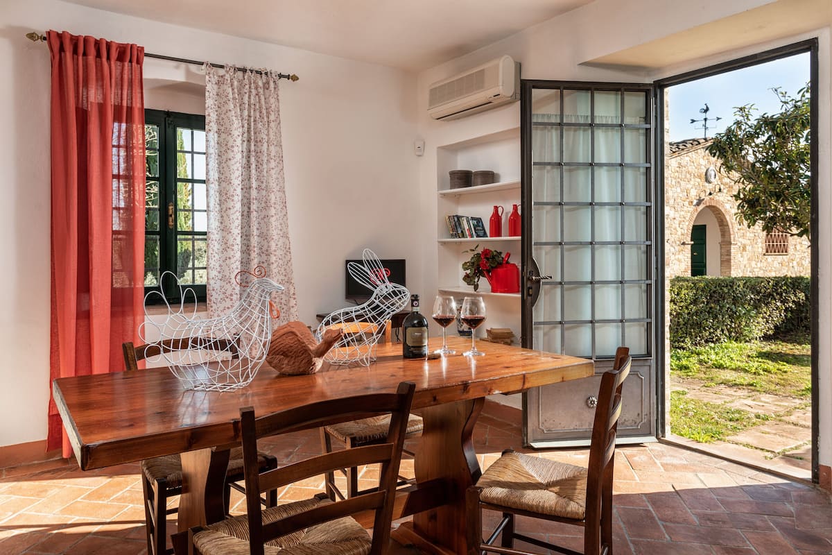 A dining area with a wooden table and chairs, a white wire basket, a wine bottle, and two wine glasses. There is a bookshelf with books and decorative items, and a view of an outdoor garden through a glass door.