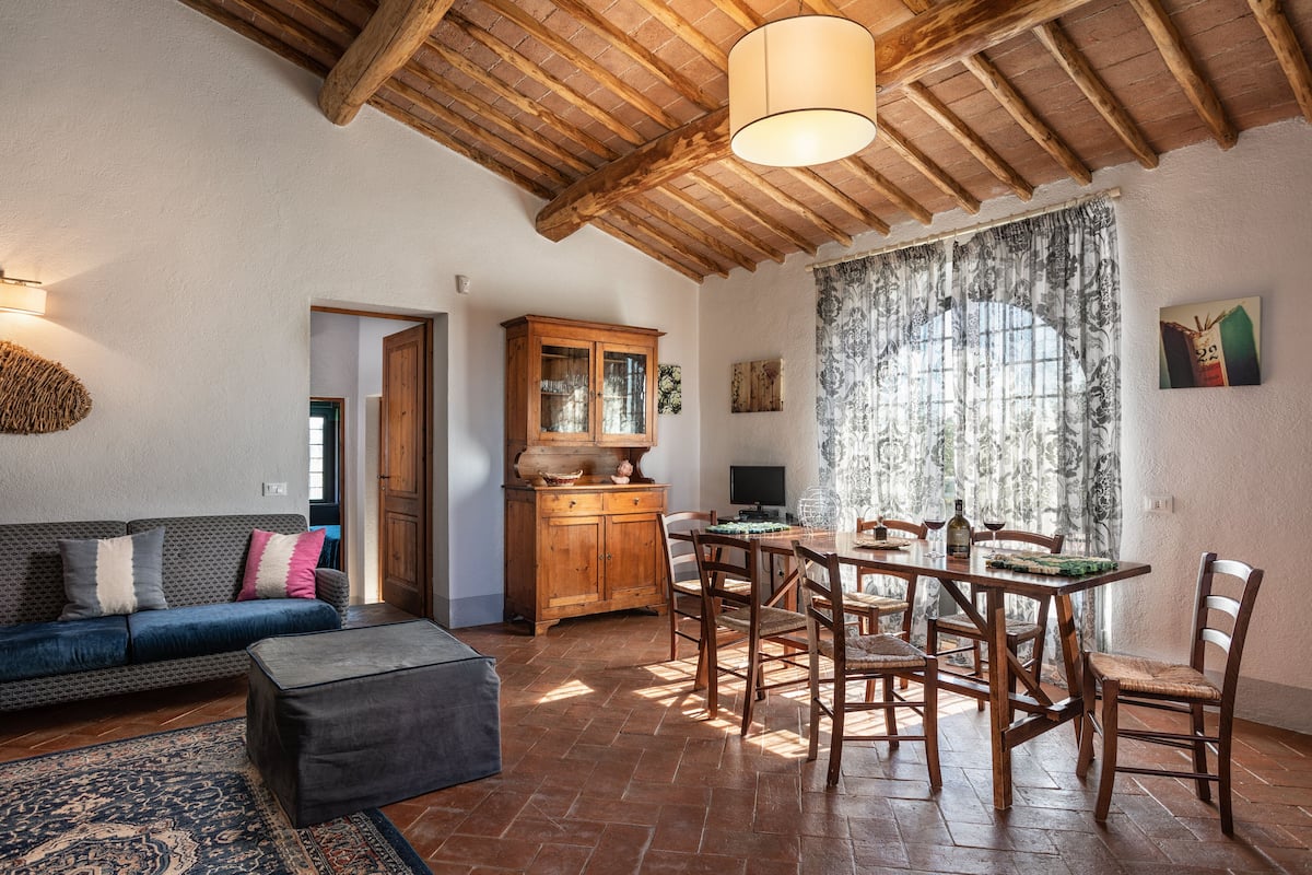 A cozy living room with a wooden ceiling, a dining area with a table and chairs, and a sofa with cushions.
