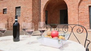 Una mesa con una botella de vino, copas y una cesta de frutas frente a un edificio de ladrillo con puertas arqueadas.