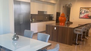 Una cocina moderna con una isla central, un comedor con mesa blanca y sillas tapizadas en azul, y una barra con taburetes.