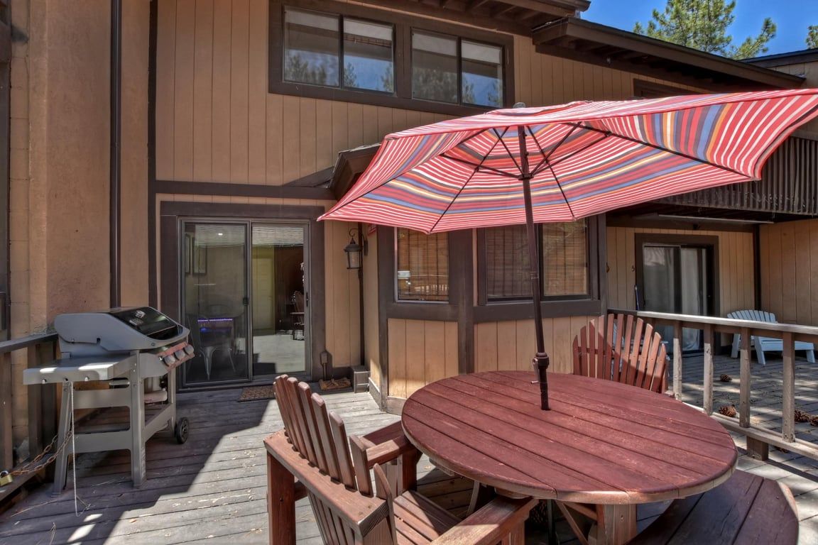 A wooden deck with a round table and chairs, a grill, and a striped umbrella.