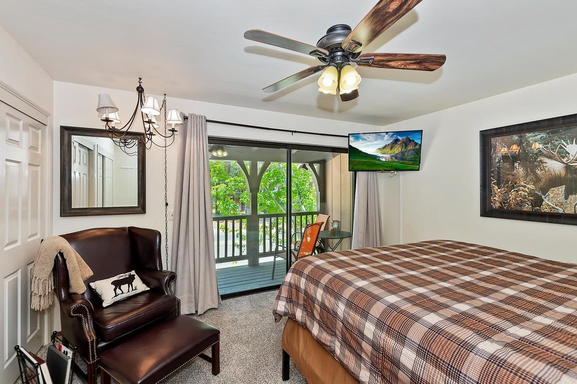 A bedroom with a bed, a chair, a ceiling fan, a television, and a balcony with a view of greenery.