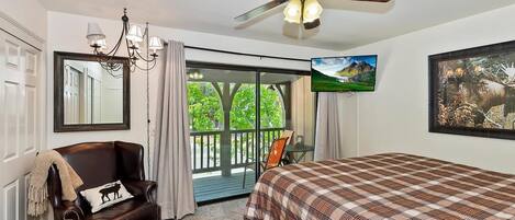 A bedroom with a bed, a chair, a ceiling fan, a television, and a balcony with a view of greenery.