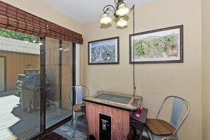 A room with a glass door leading outside, a wooden table with a game, two chairs, and framed artwork on the wall.
