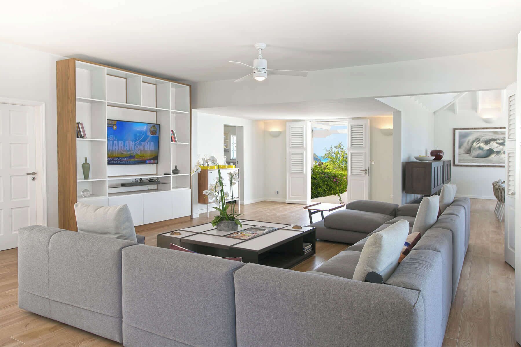 Un ampio soggiorno con un divano grigio, un tavolino da caffè e una TV su un mobile in legno. C'è una ventola a soffitto, una porta che dà su un balcone con vista e un quadro appeso al muro.