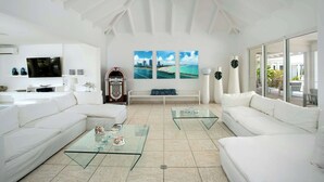 A spacious living room with white sofas, glass coffee tables, and a large TV. There are three framed pictures on the wall and a bench with a blue cushion. - Villa Encore | A Grand Caribbean Estate with Panoramic Sea Views (Philipsburg)