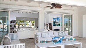 A modern living room with white furniture, a ceiling fan, and a glass door leading to a pool area. - Villa Encore | A Grand Caribbean Estate with Panoramic Sea Views (Philipsburg)