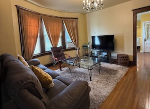 Un salon avec un canapé, un fauteuil, une table basse en verre, un téléviseur et des rideaux.