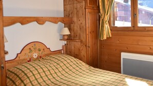 A wooden cabin room with a bed, a window with curtains, a wall-mounted lamp, and a radiator.
