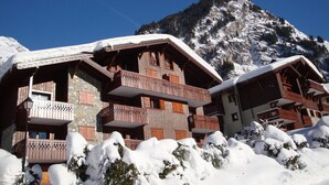 A wooden chalet with balconies, surrounded by snow-covered trees and a mountain in the background.