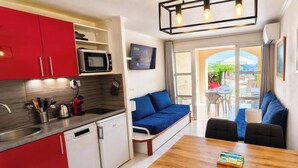 A compact kitchen with a microwave, sink, and red cabinets. A blue sofa and a wooden dining table are in the living area, with a TV mounted on the wall. - Cap Esterel le village T2 Garden level-CLIM-Sea view-Pool and parking included (Saint-Raphaël)