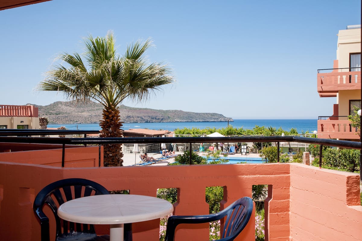A balcony with a table and chairs, overlooking a pool and a beach with a palm tree.