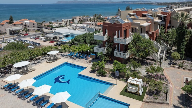 A swimming pool with a shark design, surrounded by lounge chairs and umbrellas, with a view of the sea and buildings in the background. - Ekavi Apartments (Chania)