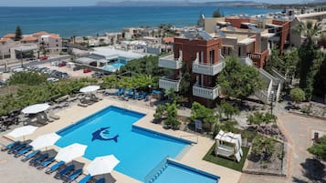 A swimming pool with a shark design, surrounded by lounge chairs and umbrellas, with a view of the sea and buildings in the background.