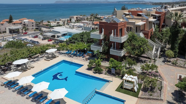 A swimming pool with a shark design, surrounded by lounge chairs and umbrellas, with a view of the sea and buildings in the background. - Ekavi Apartments (Chania)