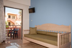 A wooden bed with a green mattress and a pillow, a TV mounted on the wall, and a balcony with chairs and a table. - Ekavi Apartments (Chania)