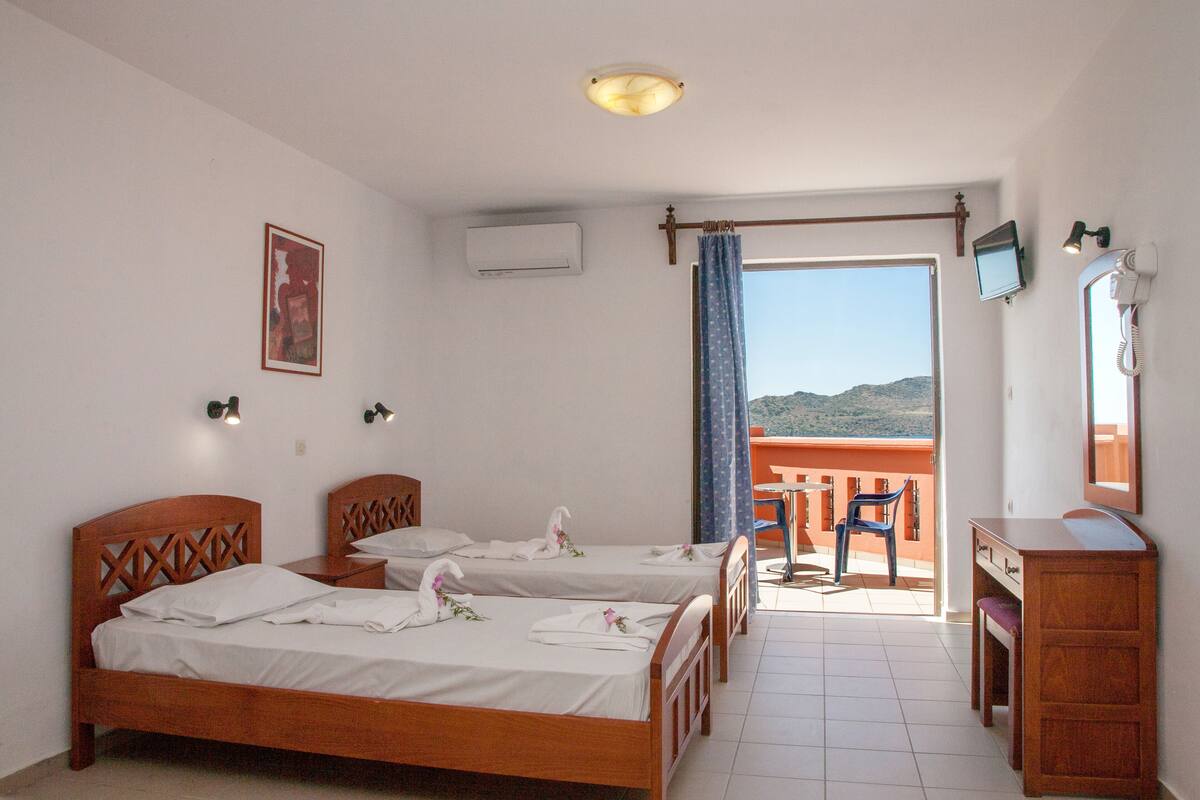 A hotel room with two wooden beds, a desk, a chair, a small table, a TV, and a balcony with a view.