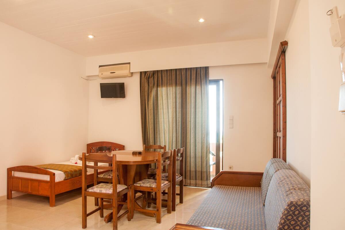 A room with a bed, a sofa, a dining table with chairs, and a TV mounted on the wall.