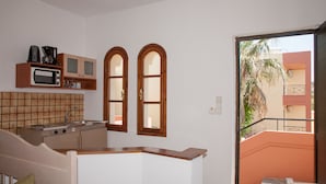 A compact kitchen with a microwave, coffee maker, and cabinets. There are two windows and a view of a building outside. - Ekavi Apartments (Chania)