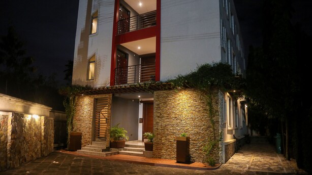 Een modern, twee verdiepingen tellend gebouw met een gevel van natuursteen, een rode balkonhek en een goed verlichte ingang met trappen en plantenbakken.