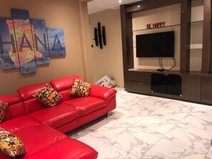 Uma sala de estar moderna com um sofá de couro vermelho, almofadas coloridas, uma televisão de tela plana e um letreiro na parede com a inscrição 'BE HAPPY'.