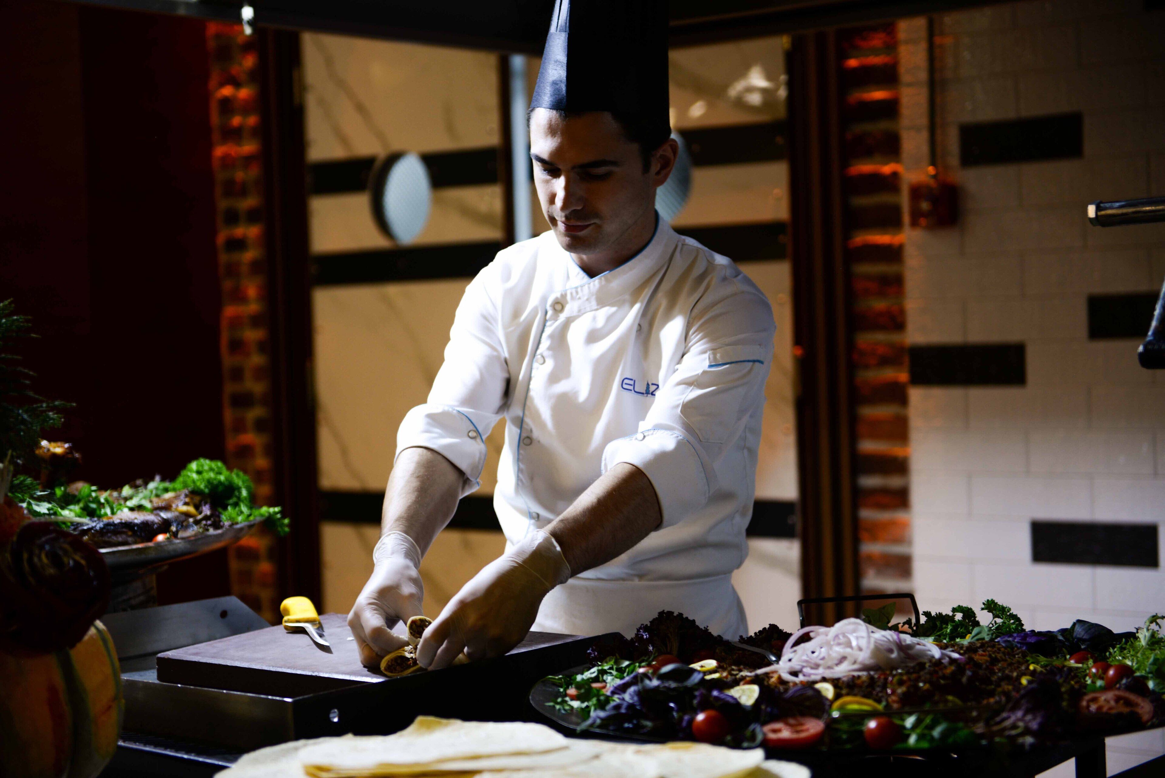 Un xef amb uniforme blanca preparant aliments al taulell, hi ha diverses verdures i una taula de tall.