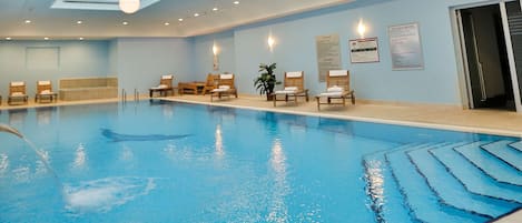 An indoor swimming pool with a skylight, lounge chairs, and a ceiling with recessed lighting.
