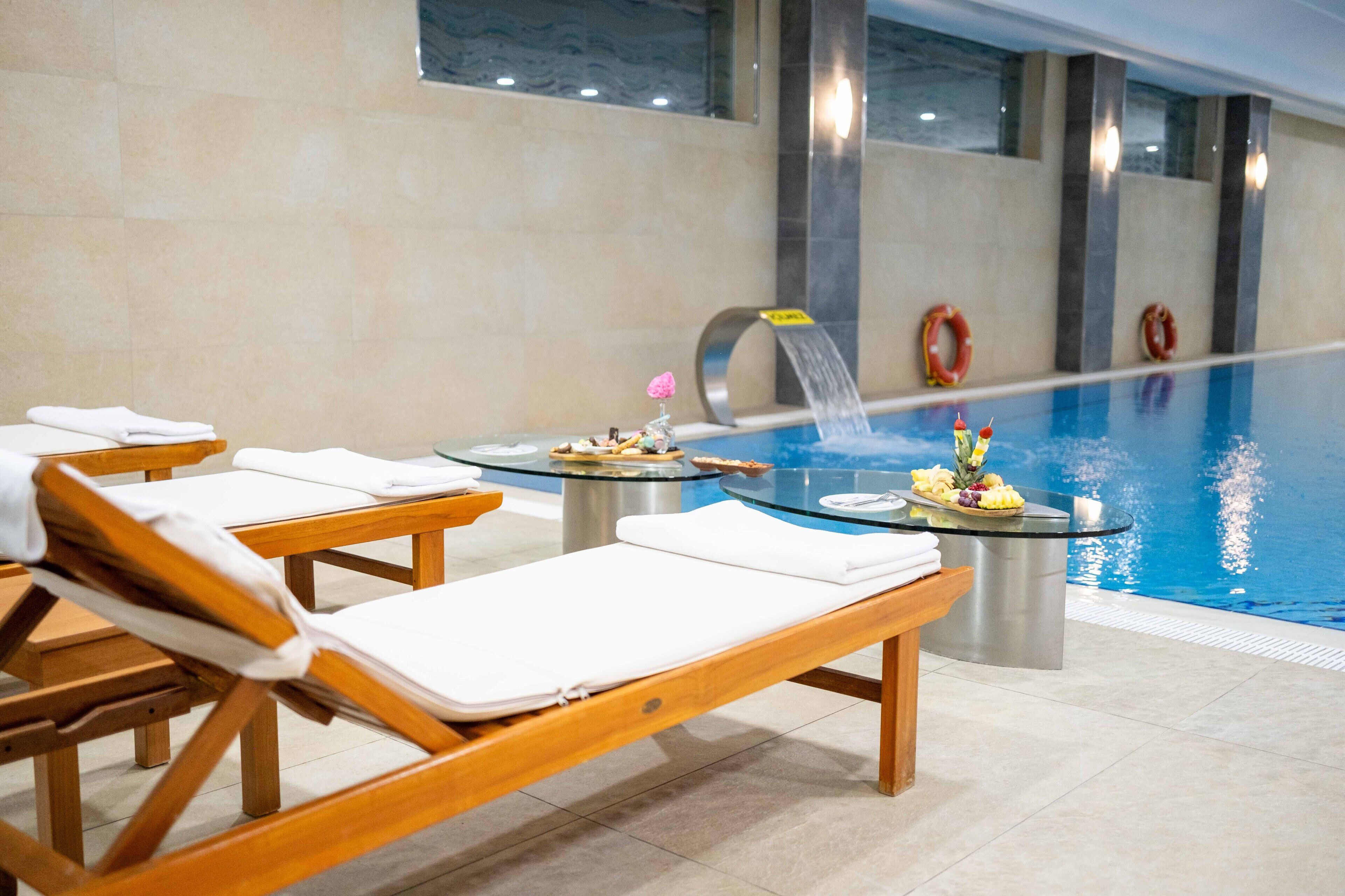 Uma área à beira da piscina com espreguiçadeiras, uma pequena mesa com frutas e flores, e uma fonte de água.