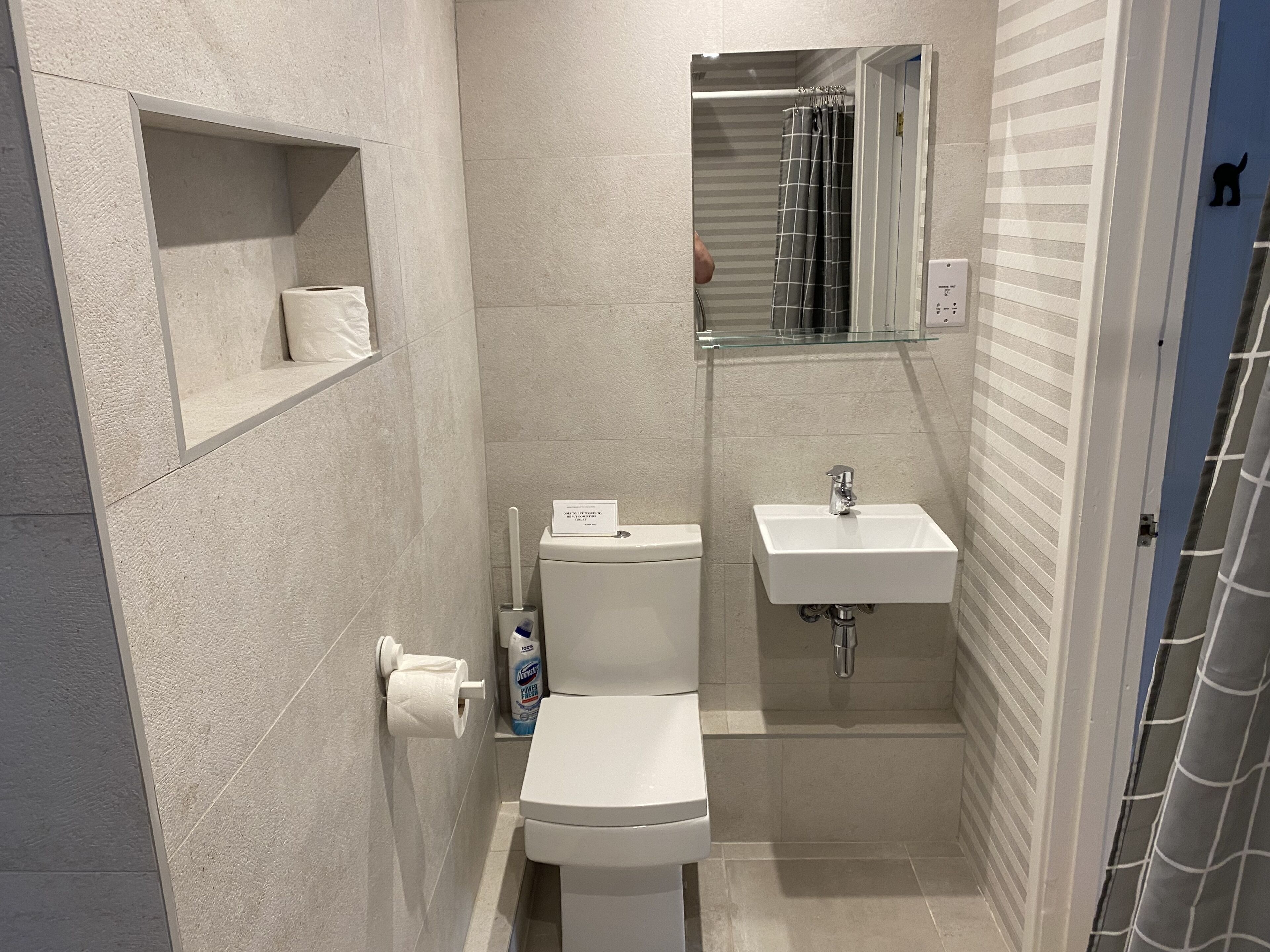 Une salle de bain moderne équipée de toilettes, d’un lavabo et d’une douche. On y trouve une étagère avec un rouleau de papier toilette et un flacon de savon pour les mains.
