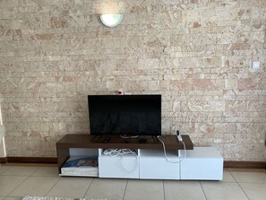 A modern living room with a flat-screen TV on a wooden entertainment unit, a stone wall, and a small white cabinet with a picture frame. - 3 bedroom penthouse with beach views (Mombasa)