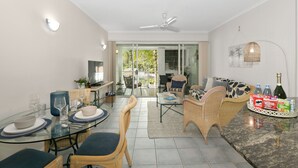 A modern living room with a glass dining table, wicker furniture, and a large window offering a view of the outdoors.