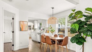 Una cocina moderna con armarios blancos, una mesa de comedor con sillas, una lámpara colgante y una planta grande en maceta.