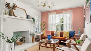 Una sala de estar con chimenea de ladrillo blanco, una mesa de centro de vidrio, un sofá con cojines de colores y una zona de comedor con mesa y sillas.