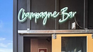 Un balcon orné d’un panneau lumineux au néon indiquant « Bar à champagne », un petit coin cuisine équipé d’un réfrigérateur, d’un évier et d’un plan de travail agrémenté d’ustensiles de bar.