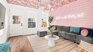 Un salon avec un plafond orné de motifs floraux, un canapé gris, une table basse blanche et un mur rose agrémenté d'un néon.
