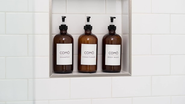 Três frascos de produtos COMÓ numa prateleira de azulejos brancos.