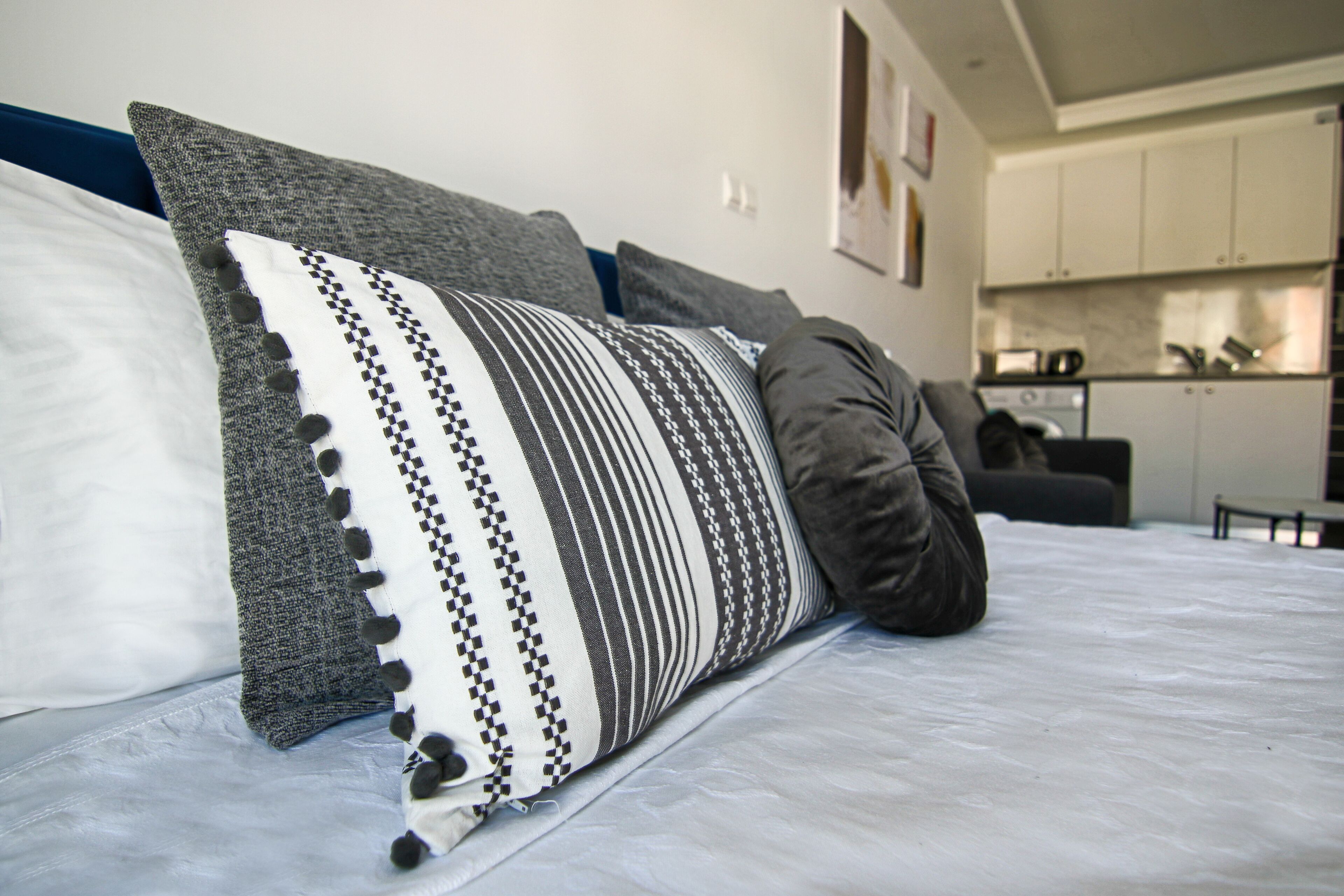 Een bed met gekleurde kussens en een grijze deken in een kamer, met een keuken op de achtergrond.