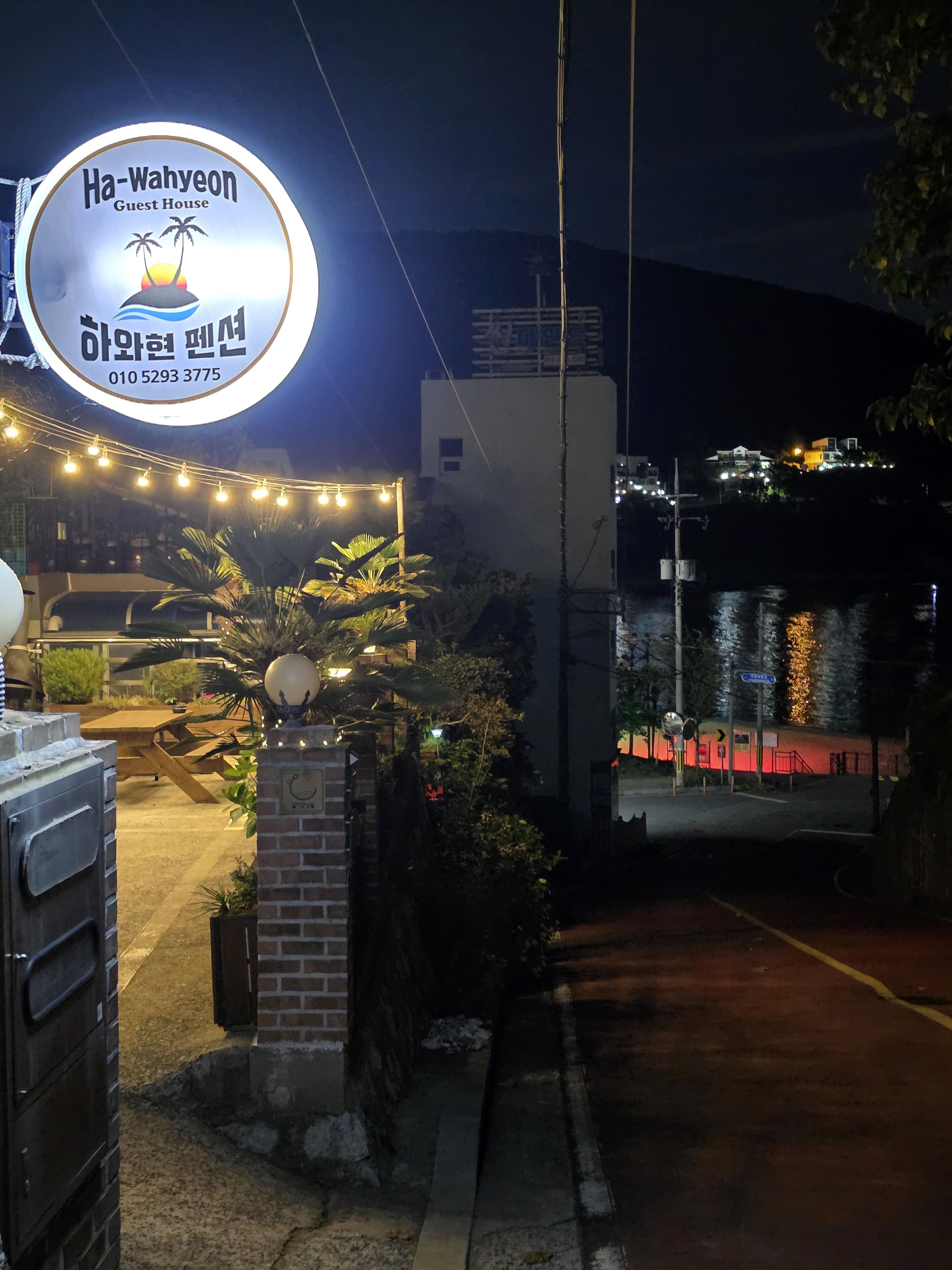 A guest house sign with palm trees and a sunset, a brick entrance, and a street view at night.