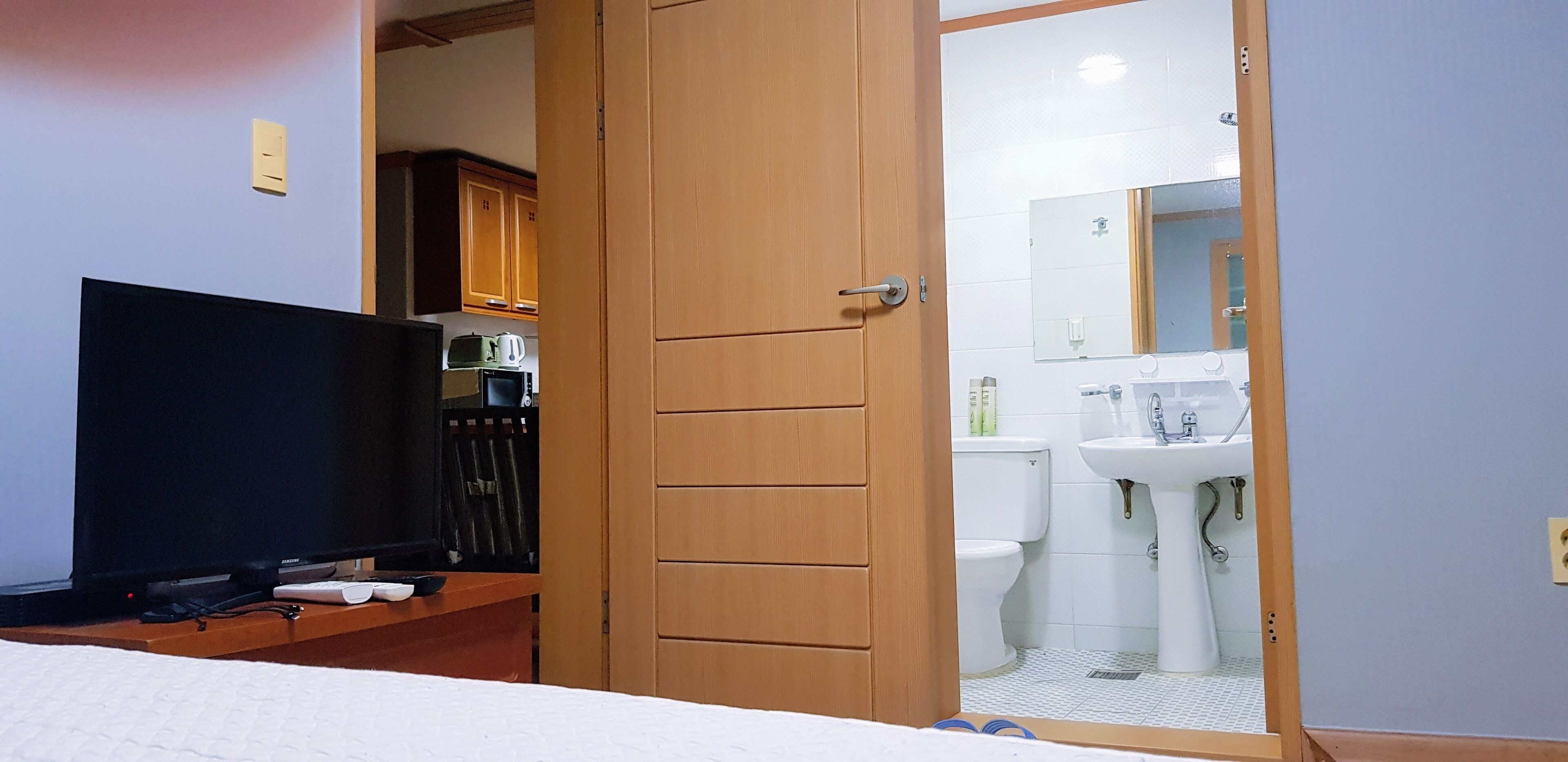 Une chambre avec une télévision, une porte en bois et une vue sur une salle de bain équipée d’un lavabo et de toilettes.